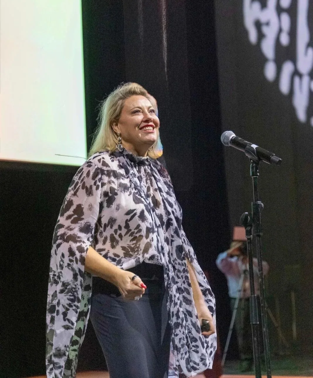 Kylie Ward smiling on stage, standing near a microphone, wearing a black and white animal print blouse and dark pants.