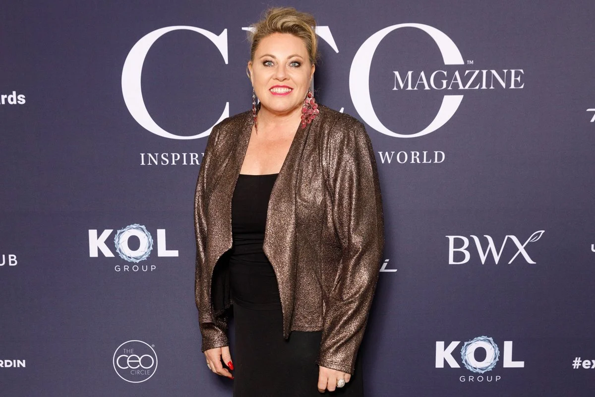 Kylie Ward with blonde hair in an updo, wearing a metallic brown jacket over a black dress, standing in front of a dark backdrop with white text and logos for CEO Magazine