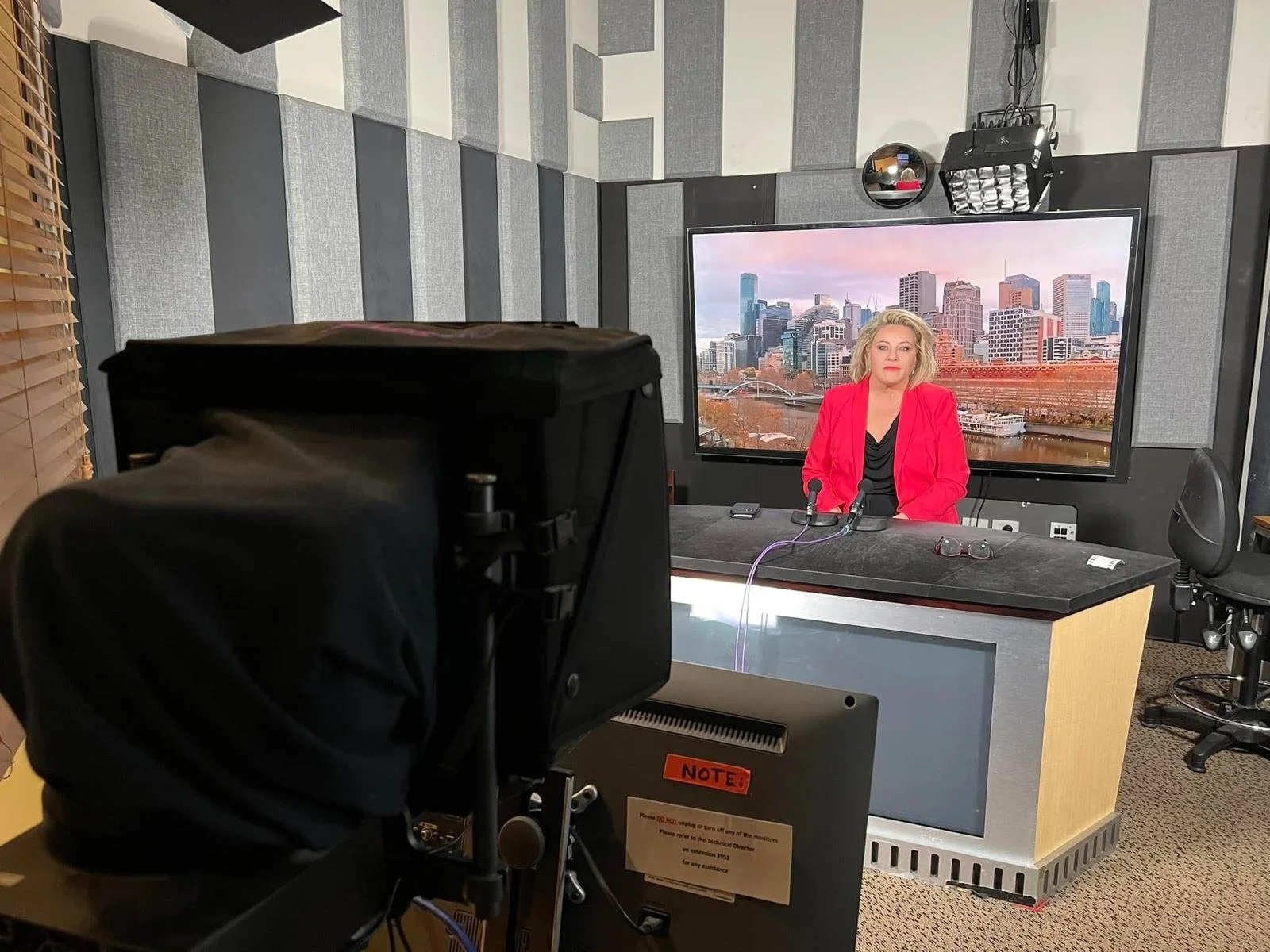 Kylie Ward in a red blazer sitting at a news desk with a cityscape background on a large screen behind her, in a recording studio.