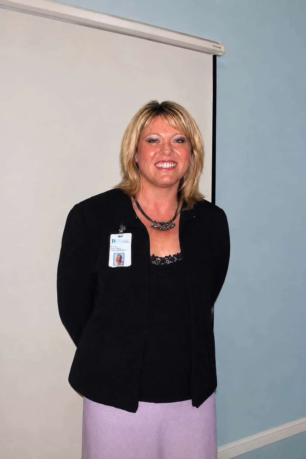 A smiling woman, Kylie Ward, with blonde hair wearing a black blazer, lavender skirt, and an ID badge, standing indoors against a background of a white wall and a light blue wall.
