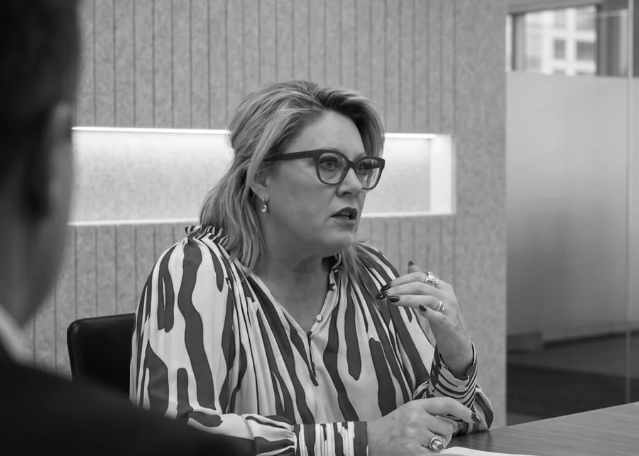Kylie Ward with glasses and shoulder-length hair, wearing a zebra-print blouse, is sitting at a table, engaged in conversation in a modern office setting.