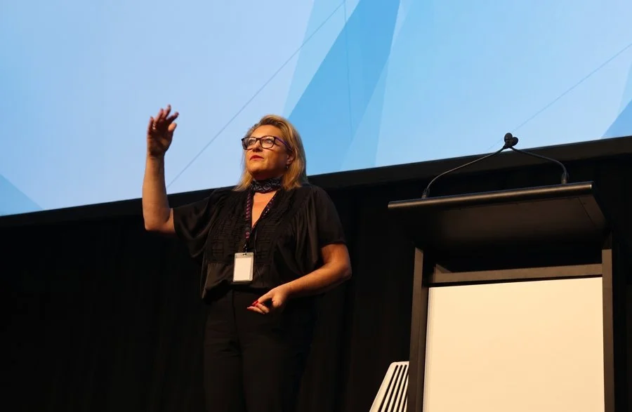 Kylie Ward presenting in front of a large blue projector screen standing next to a podium, gesturing as she speaks to the audience