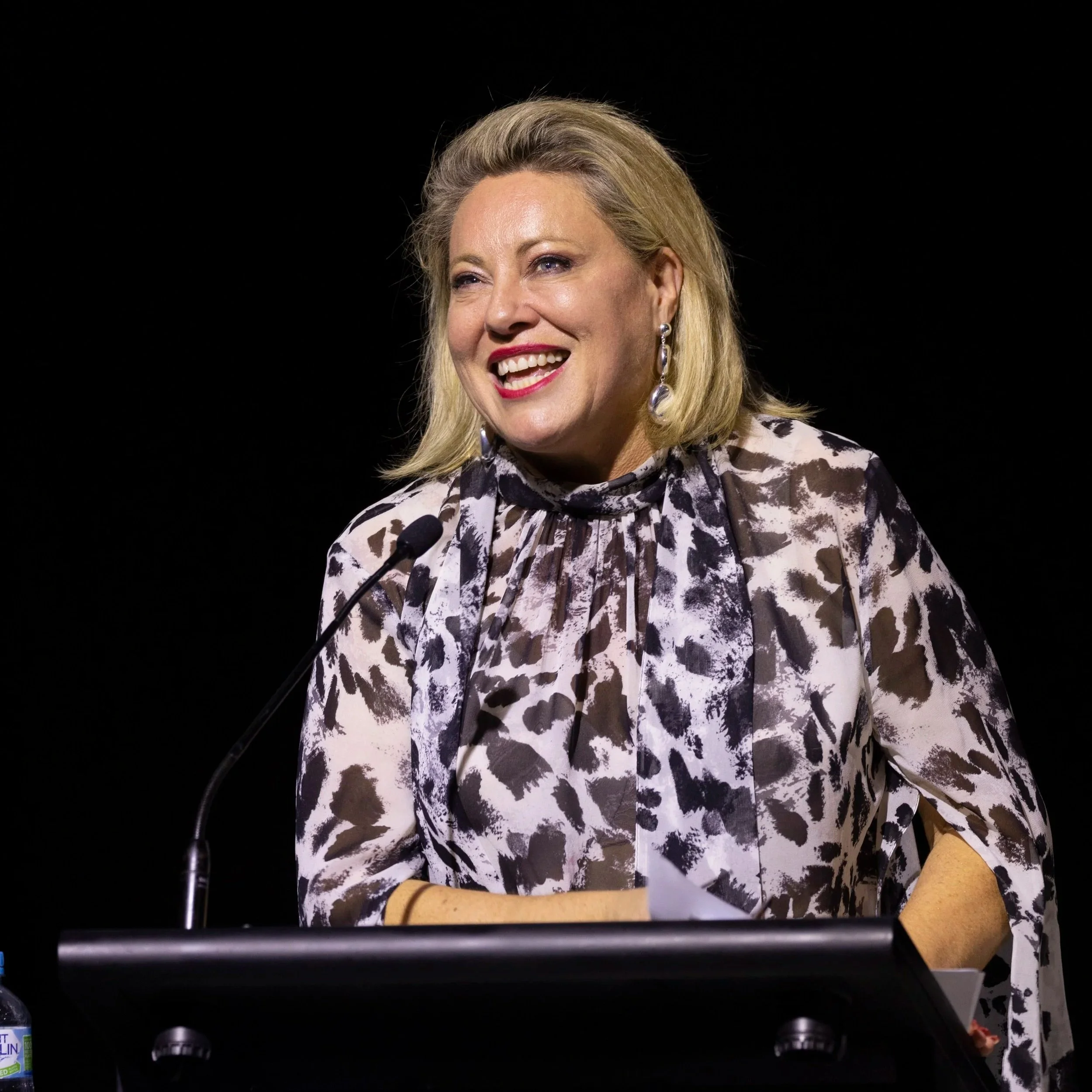 Kylie Ward smiling on stage while speaking, wearing a patterned blouse, with a microphone in front of her.