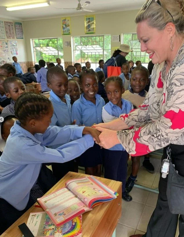 Kylie Ward shaking hands with a young girl in a classroom full of students dressed in blue school uniforms, with smiling classmates watching