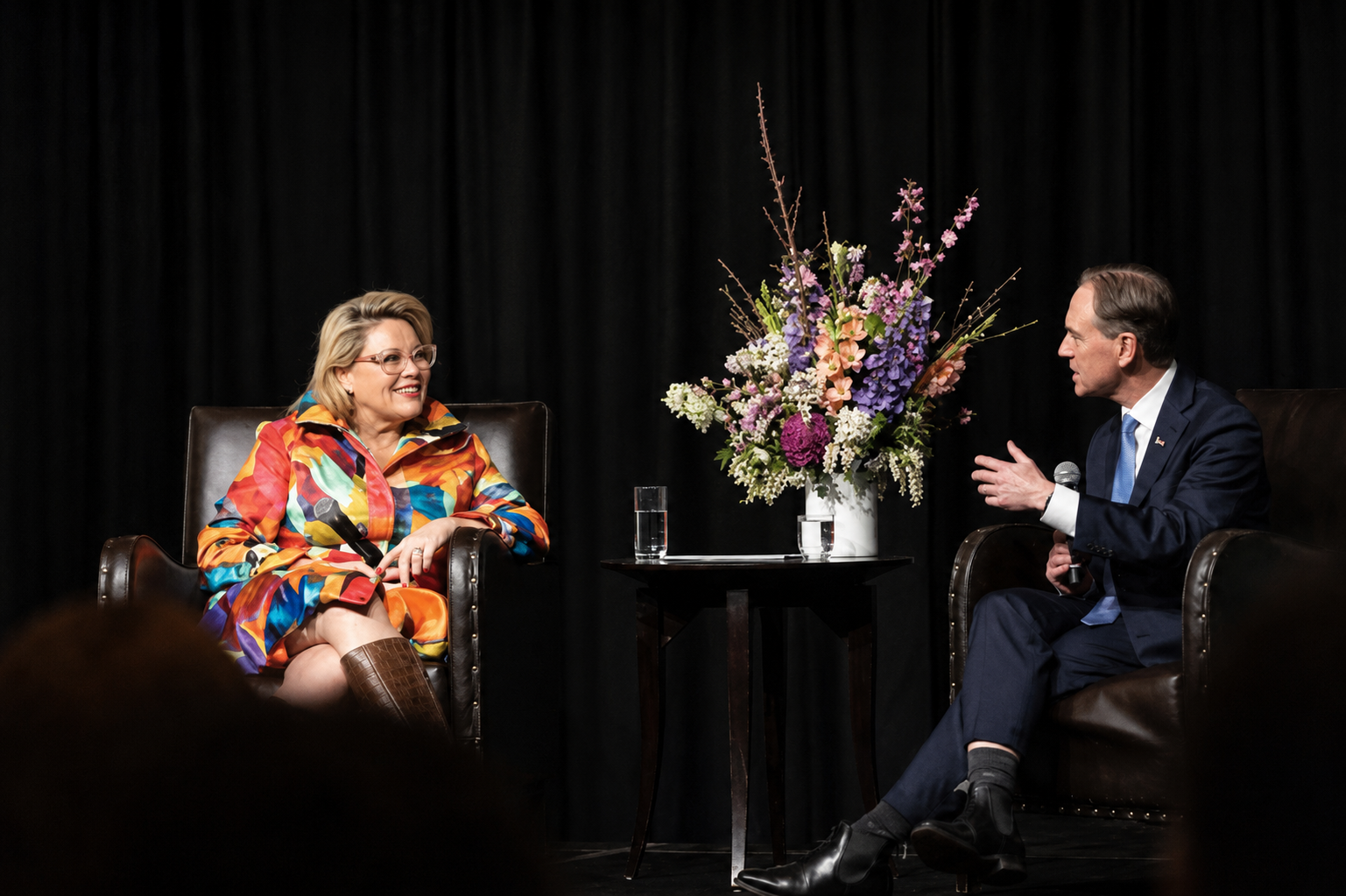 Kylie Ward in a colourful long sleeve outfit sitting in a lounge chair in stage opposite a male also sitting with a microphone asking Kylie a question with a large bouquet of flowers between them
