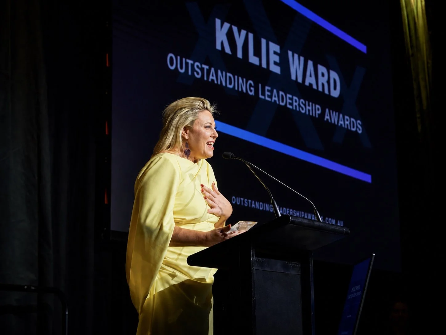 Kylie Ward speaking at a podium during the Outstanding Leadership Awards, wearing a yellow outfit, with a large display screen behind her showing event details.