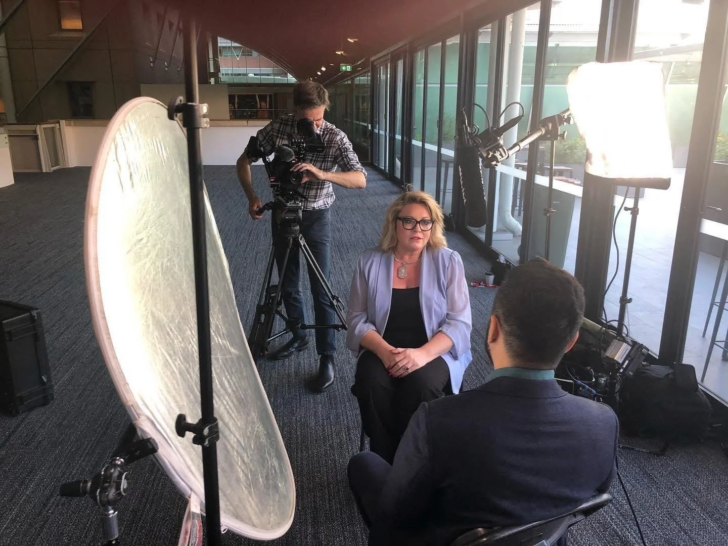 Behind the scenes of a video shoot, with Kylie Ward seated for an interview as a cameraman films from an office corridor.