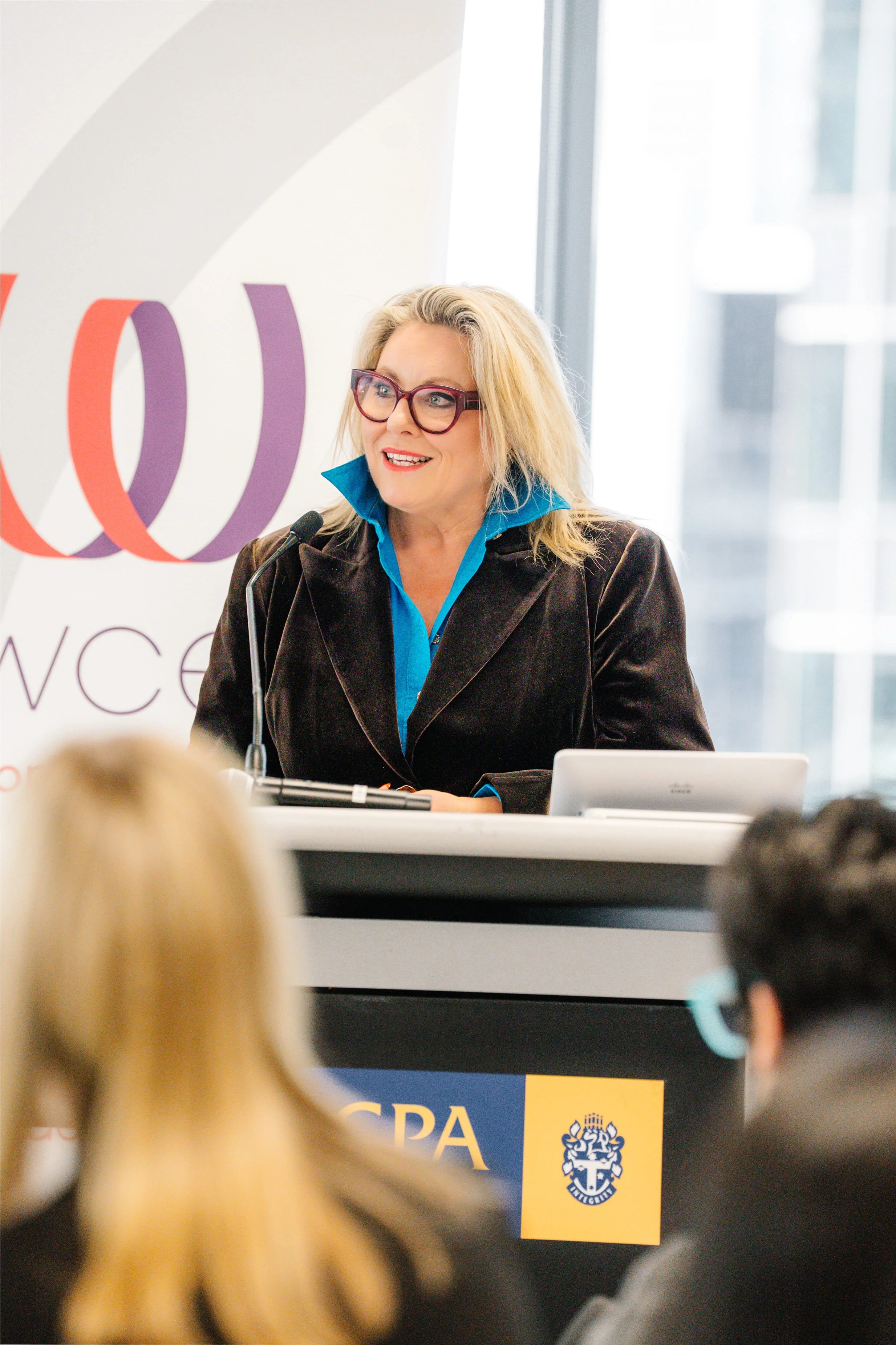 Kylie Ward wearing glasses and a dark brown jacket, speaking at a podium during a conference. Behind her is a large white and purple banner and another banner with blue and yellow colours and the text "CPA" and a logo.