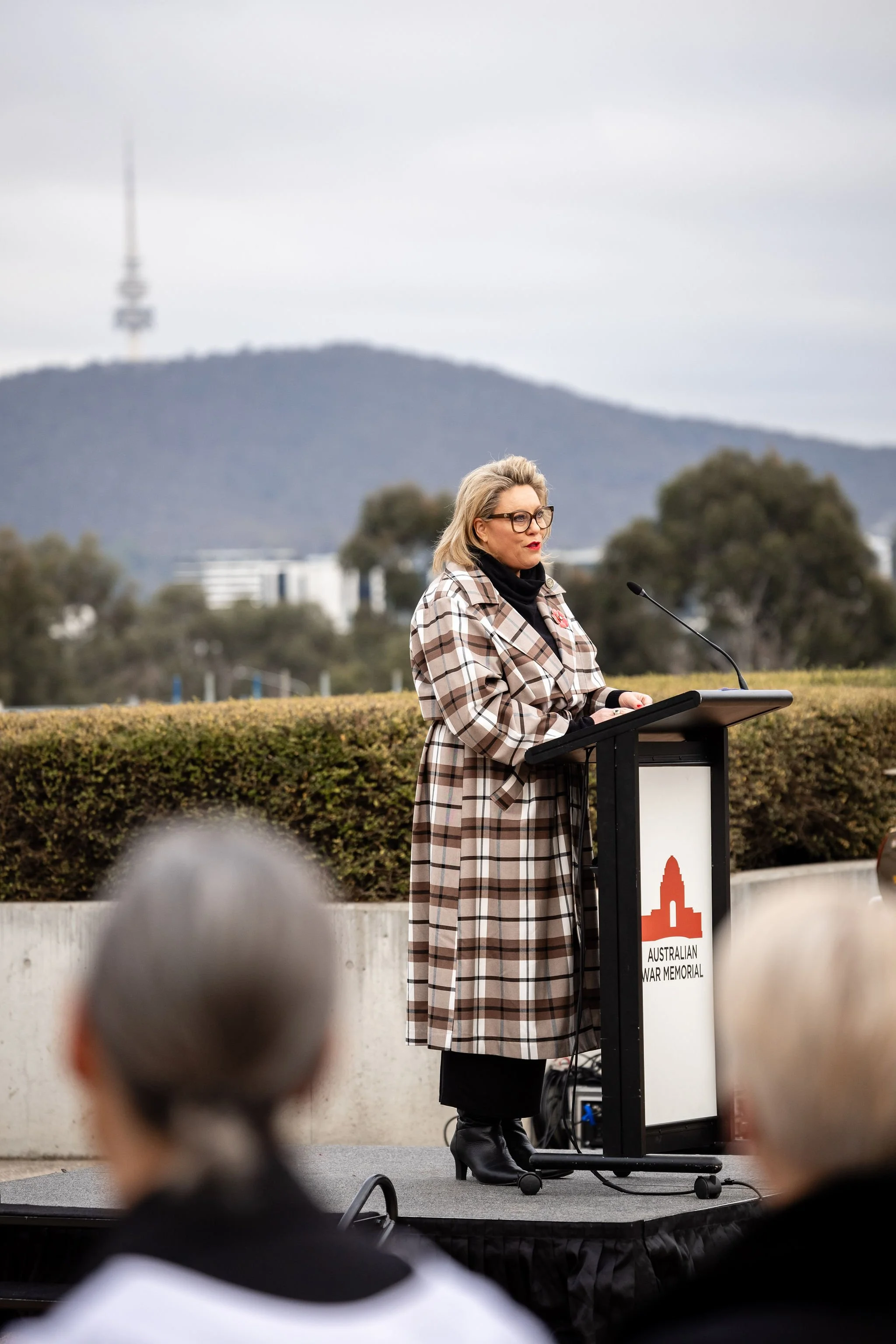 Kylie Ward speaking at an outdoor podium at the Australian War Memorial, with Canberra's Black Mountain Tower visible in the background.