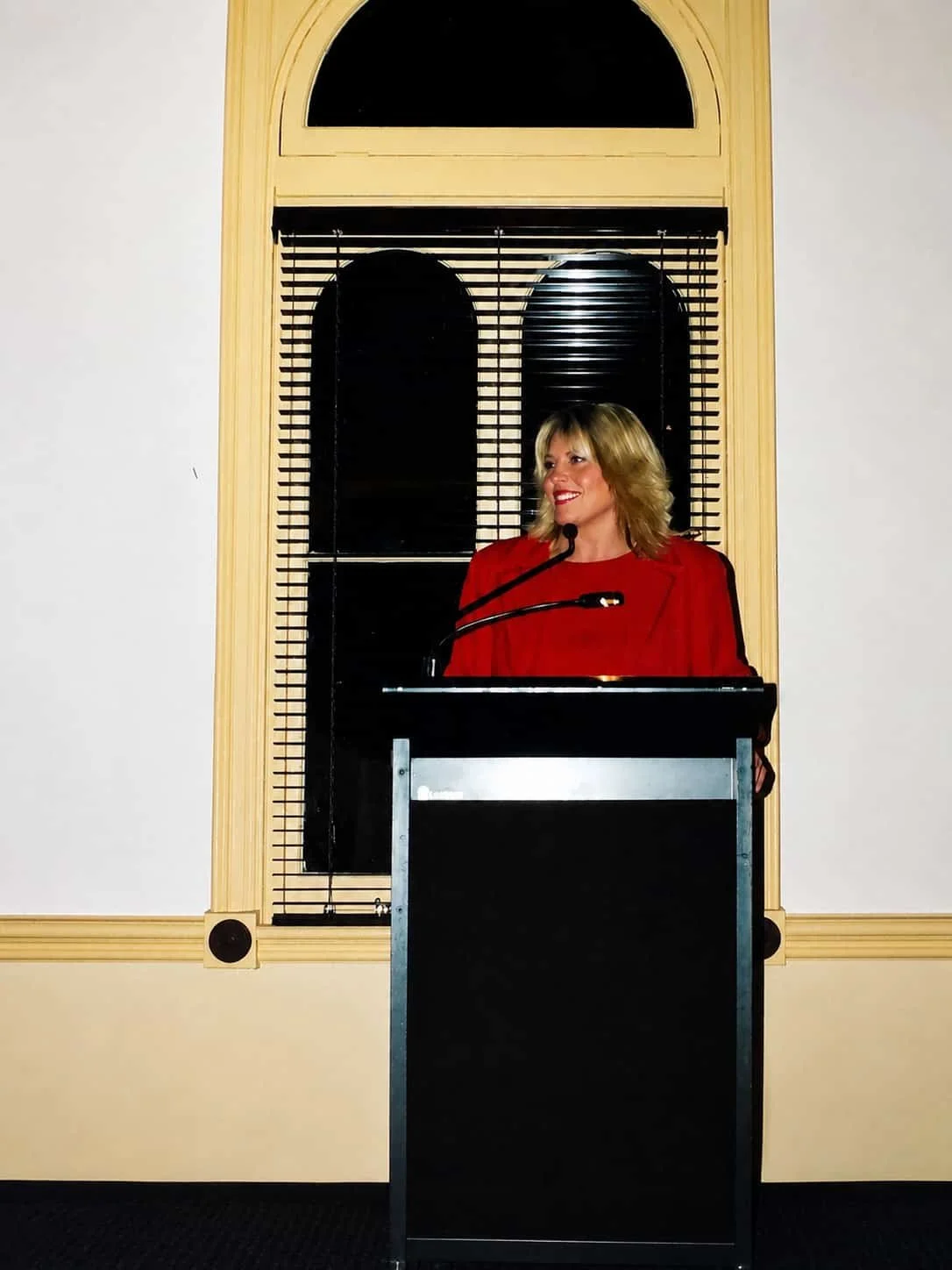 Kylie Ward in a red blazer speaking at a podium in front of a window with black blinds.