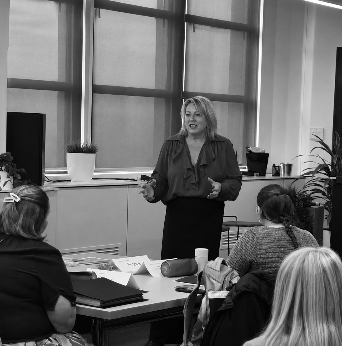 Kylie Ward mid speech wearing a blouse and dark pants, speaking to a room of professionals sitting at tables in a modern office setting and large windows behind her