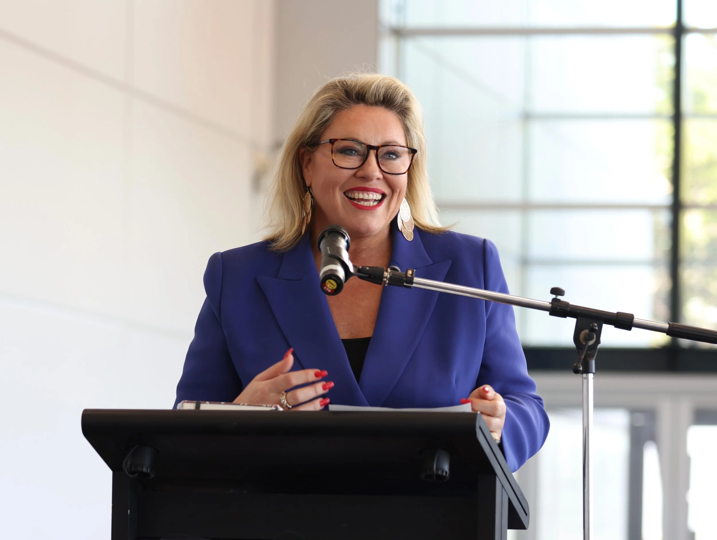 Kylie Ward with blonde hair, glasses, and earrings, wearing a blue blazer, speaking at a podium with a microphone in front of her. She is smiling and gesturing with her right hand. The background shows large windows with natural light.