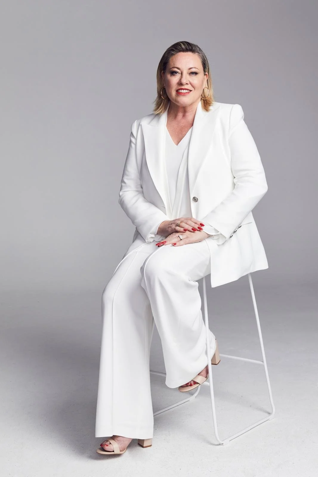Kylie Ward with shoulder-length hair, wearing a white suit with a blazer and wide-legged trousers, seated on a white chair against a plain gray background.