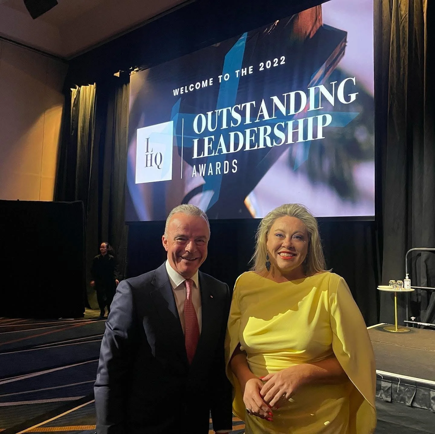 Kylie Ward, wearing a yellow dress, stands with Brendan Nelson at the 2022 Outstanding Leadership Awards, with the event signage behind them.