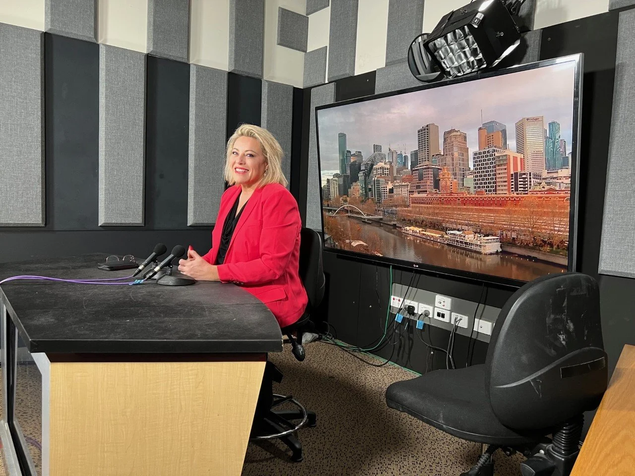 Kylie Ward in a red blazer sitting at a press conference table with multiple microphones, in a tv studio with soundproofing panels, and a large screen displaying the Melbourne city skyline and river behind her.
