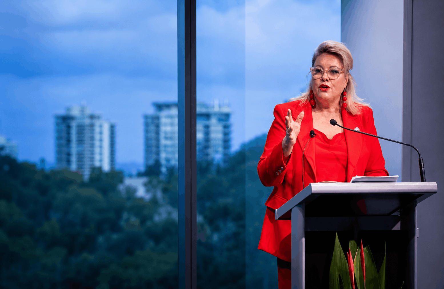 Kylie Ward in a red blazer, speaking behind a microphone on a podium with a large window behind looking out to a city scape