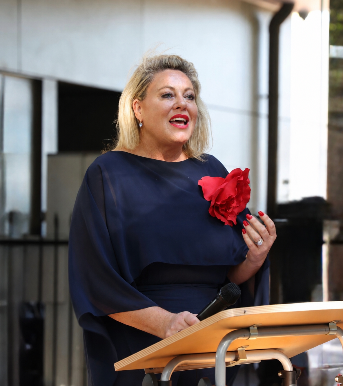 Kylie Ward with blonde hair, wearing a dark blue dress with a large red flower on her chest, holding a microphone and speaking at a podium.