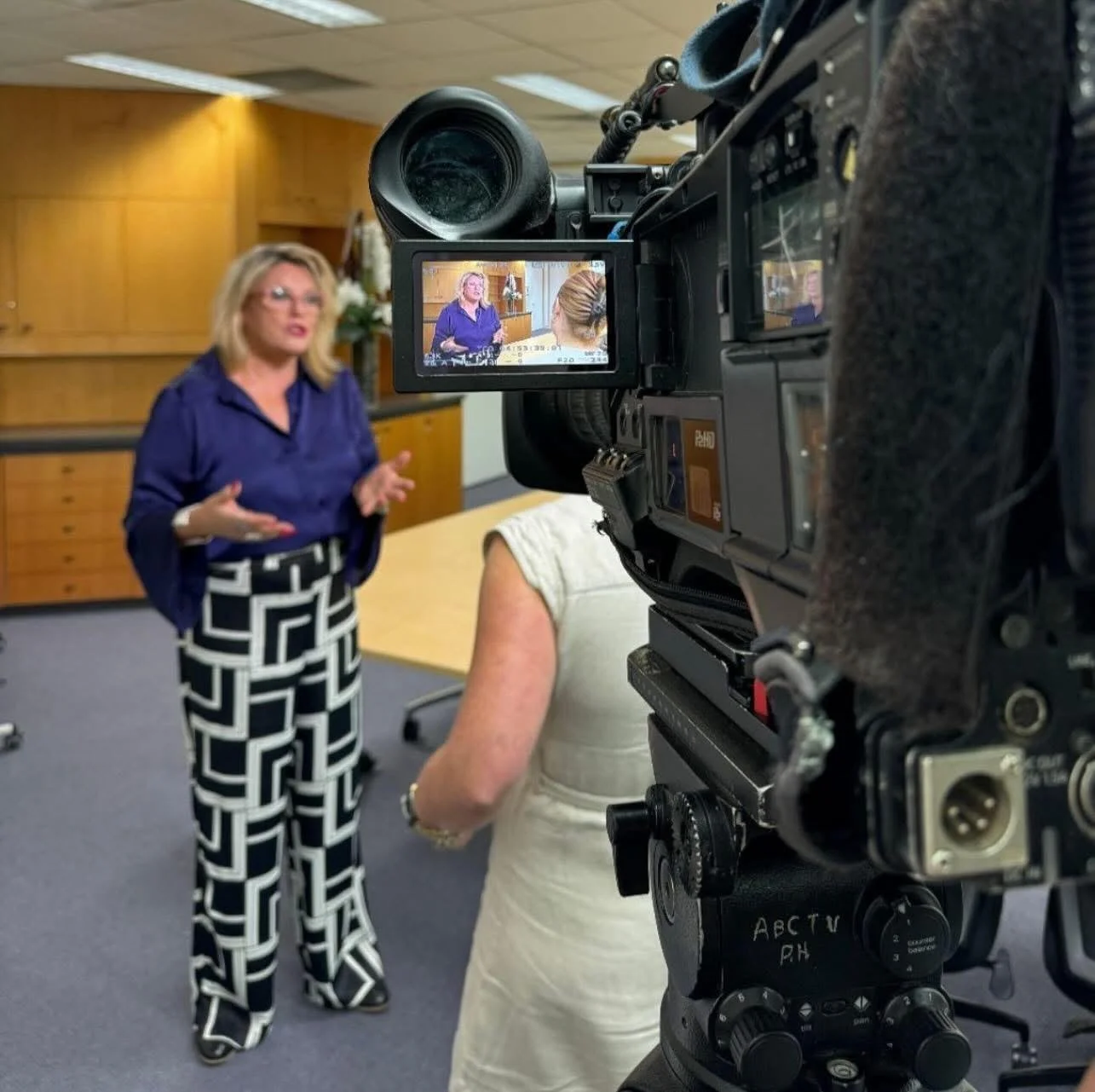 Kylie Ward in a purple shirt and black-and-white patterned pants is being filmed in an indoor setting. A camera captures her speaking to an interviewer, with the camera's display showing her face and gestures.