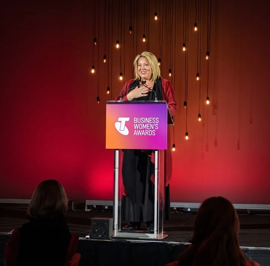 Kylie Ward behind a podium with a sign that says 'Telstra Business Women's Awards', smiling and speaking after winning an award, with hanging light bulbs on a red background.