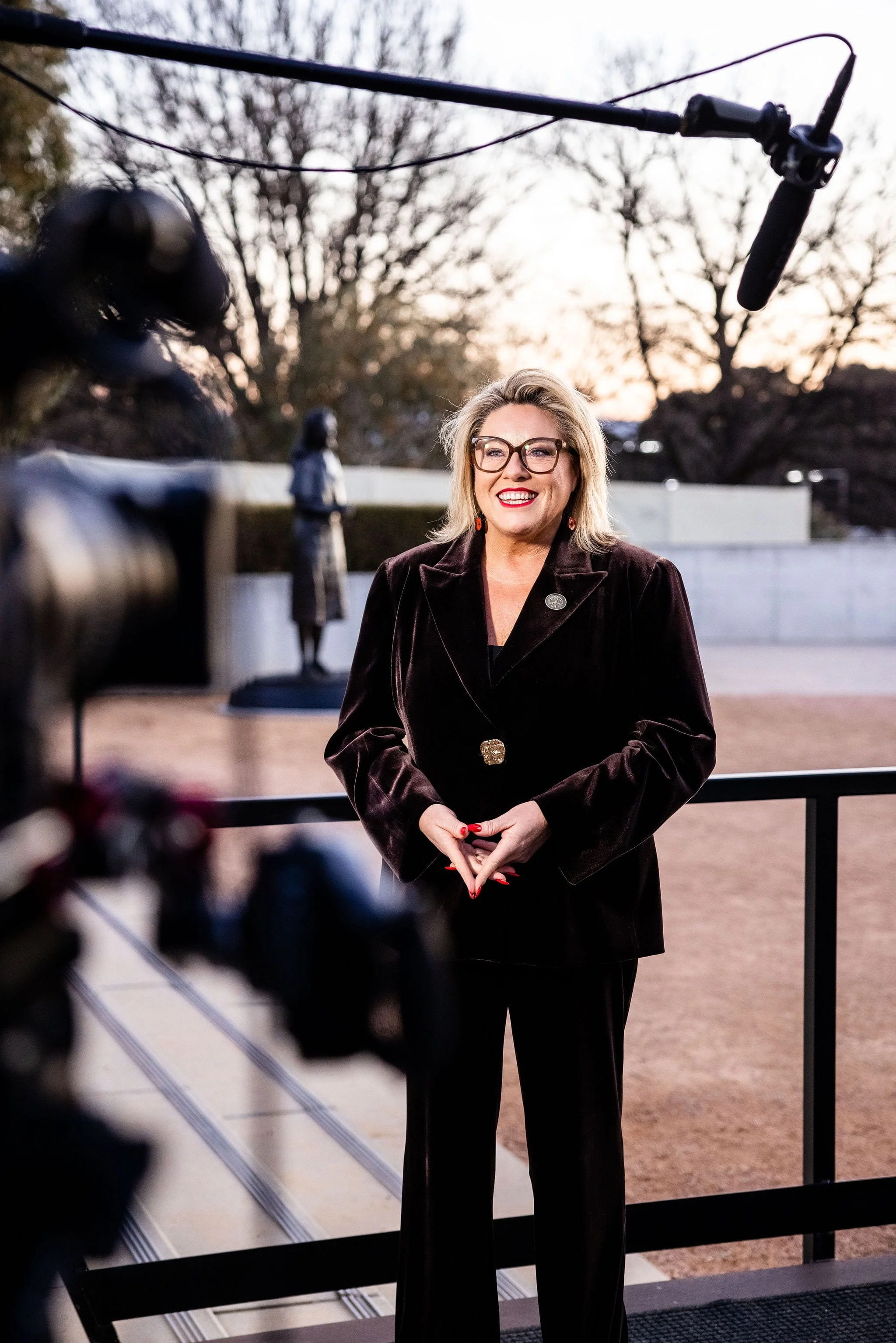 Kylie Ward with blonde hair, glasses, and red lipstick standing outdoors during sunset, dressed in a black velvet blazer and black pants, being filmed with a professional camera.