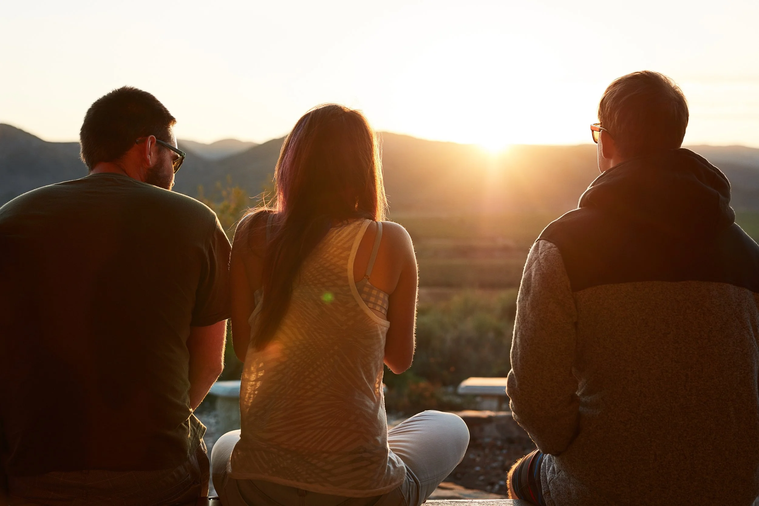 Three people sitting outdoors during sunset, facing the horizon with mountains in the background.