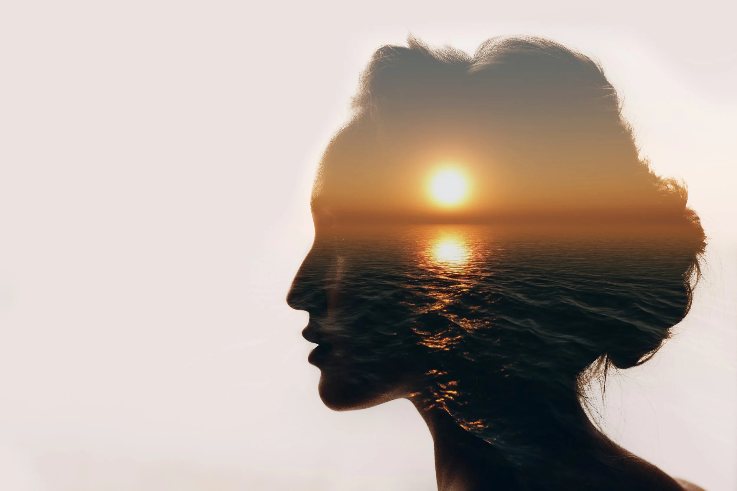 A double exposure image of a woman's profile with a sunset over the ocean inside her silhouette.
