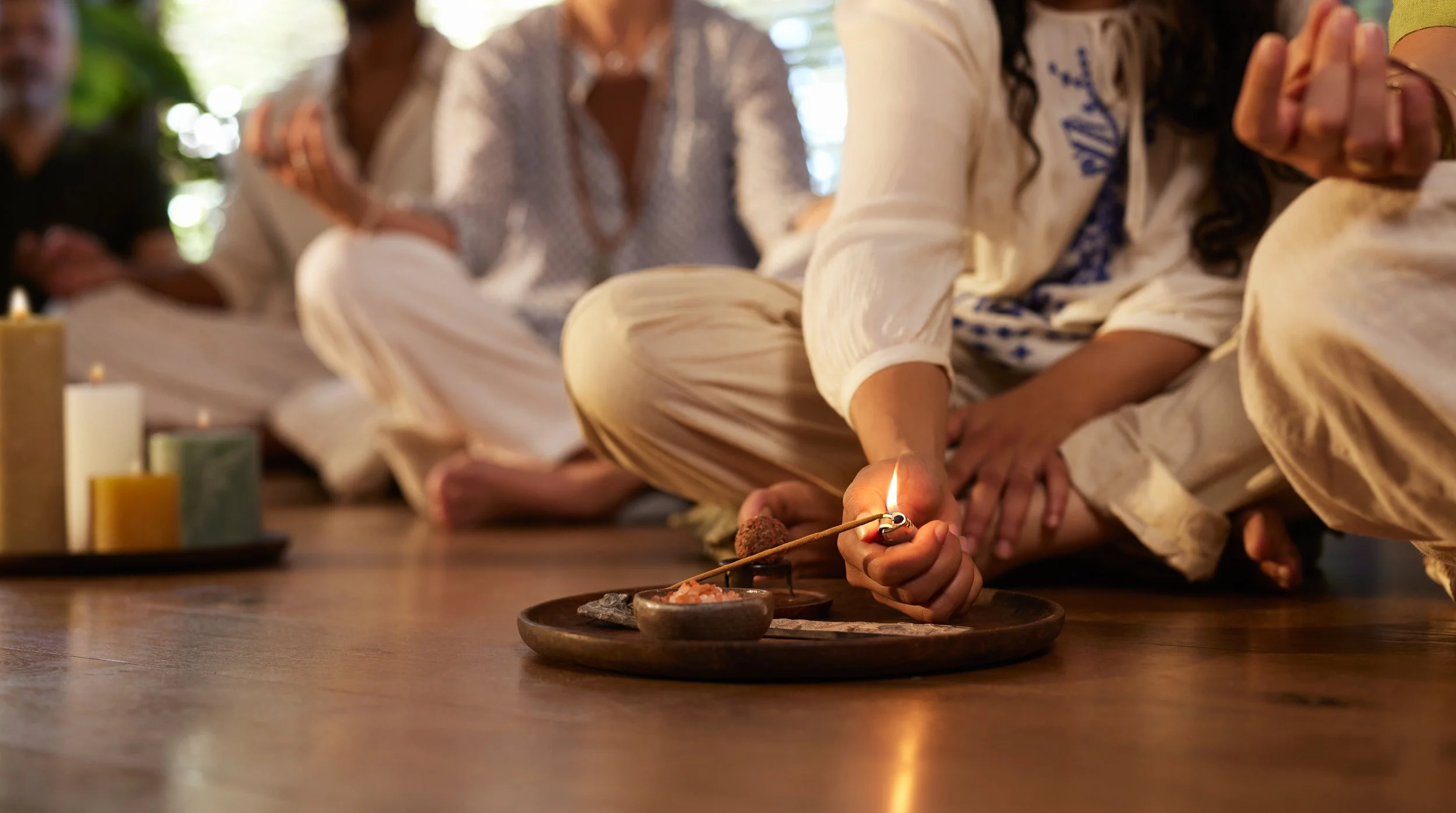 Group of people sitting cross-legged on the floor during a ceremony, with candles and small bowls in front of them. One person is holding a lit match near a candle.