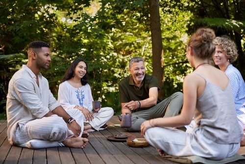Group of five people sitting on a wooden deck outdoors, smiling and engaging in conversation among trees.