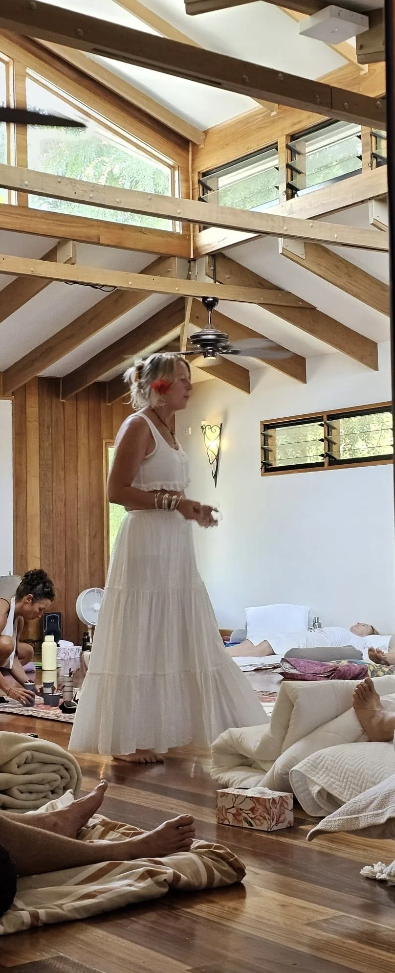 A woman in a white dress walking barefoot in a brightly lit room with wooden walls and ceiling beams. Several people are lying on the floor and on cushions, and there are blankets, a tissue box, and a small fan in the background. Outside the windows, green trees are visible.