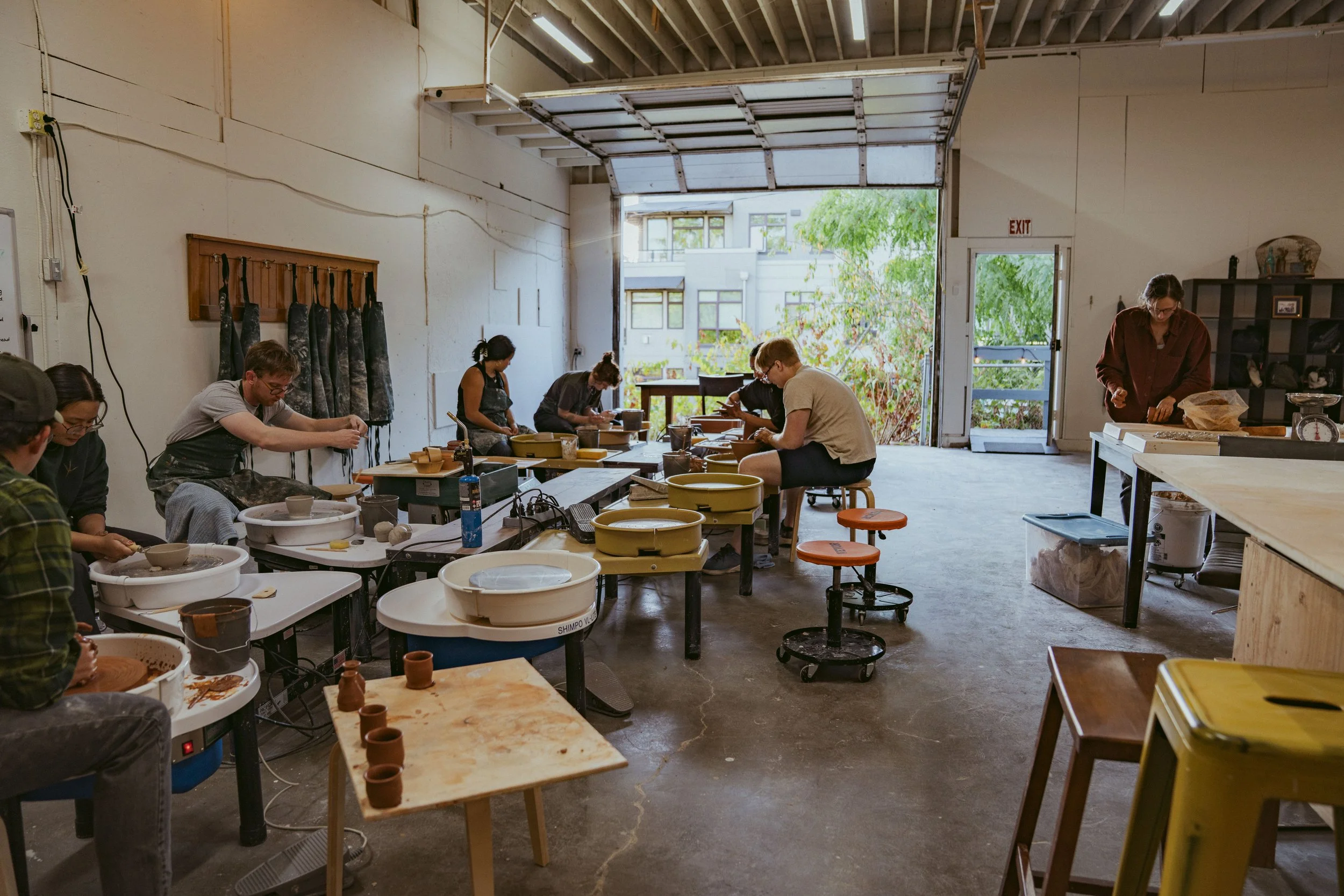 People throwing clay on pottery wheels inside a pottery studio with large open garage door and trees outside.