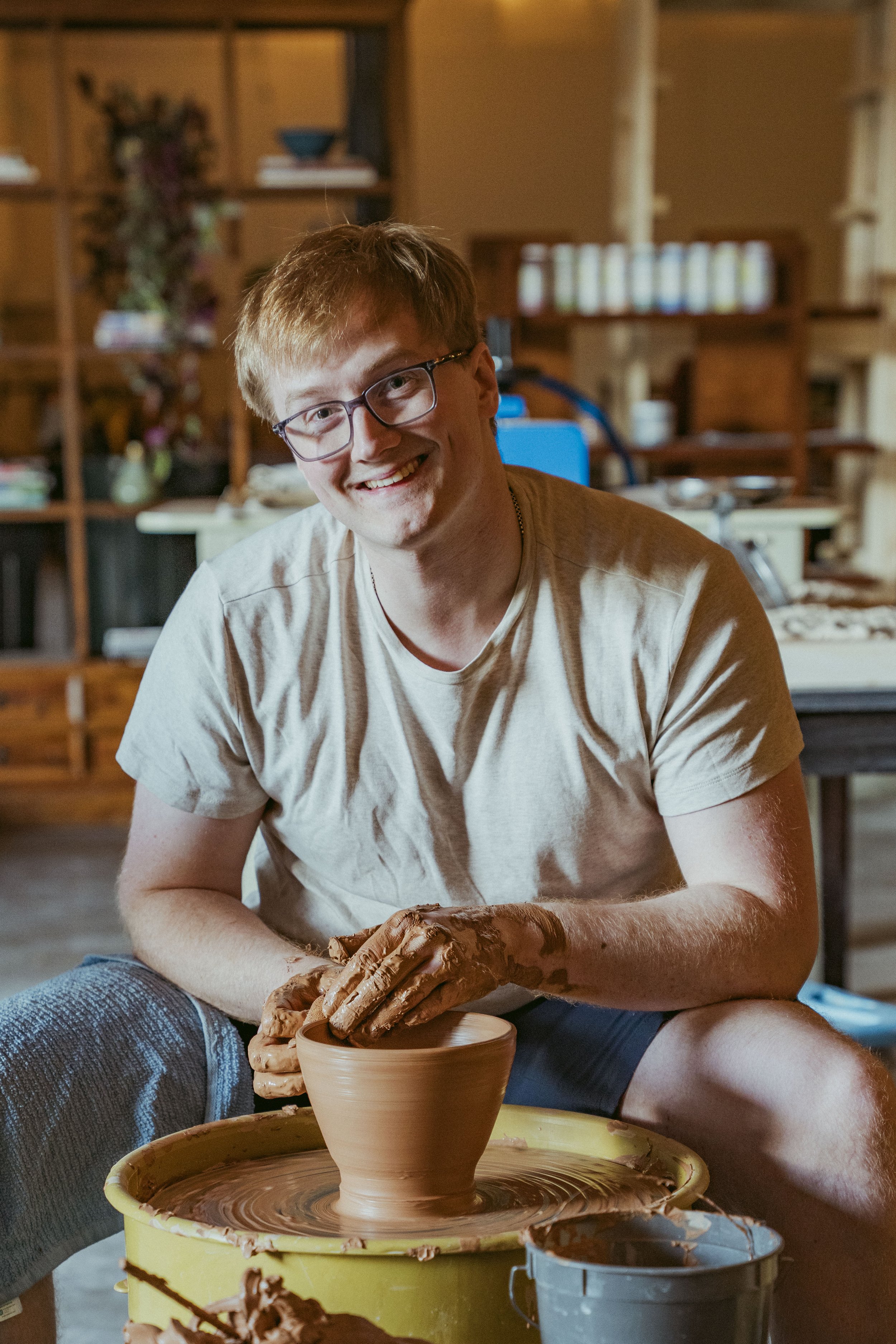 A young man with glasses and a white t-shirt is smiling while working with clay on a pottery wheel.