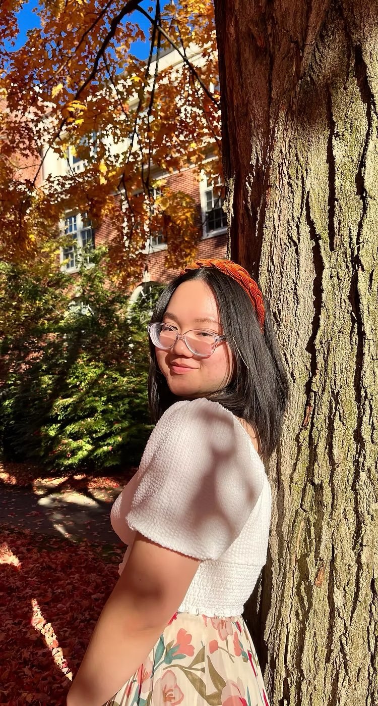 Young woman with black hair, glasses, and a floral skirt leaning against a tree with autumn leaves in the background, daytime