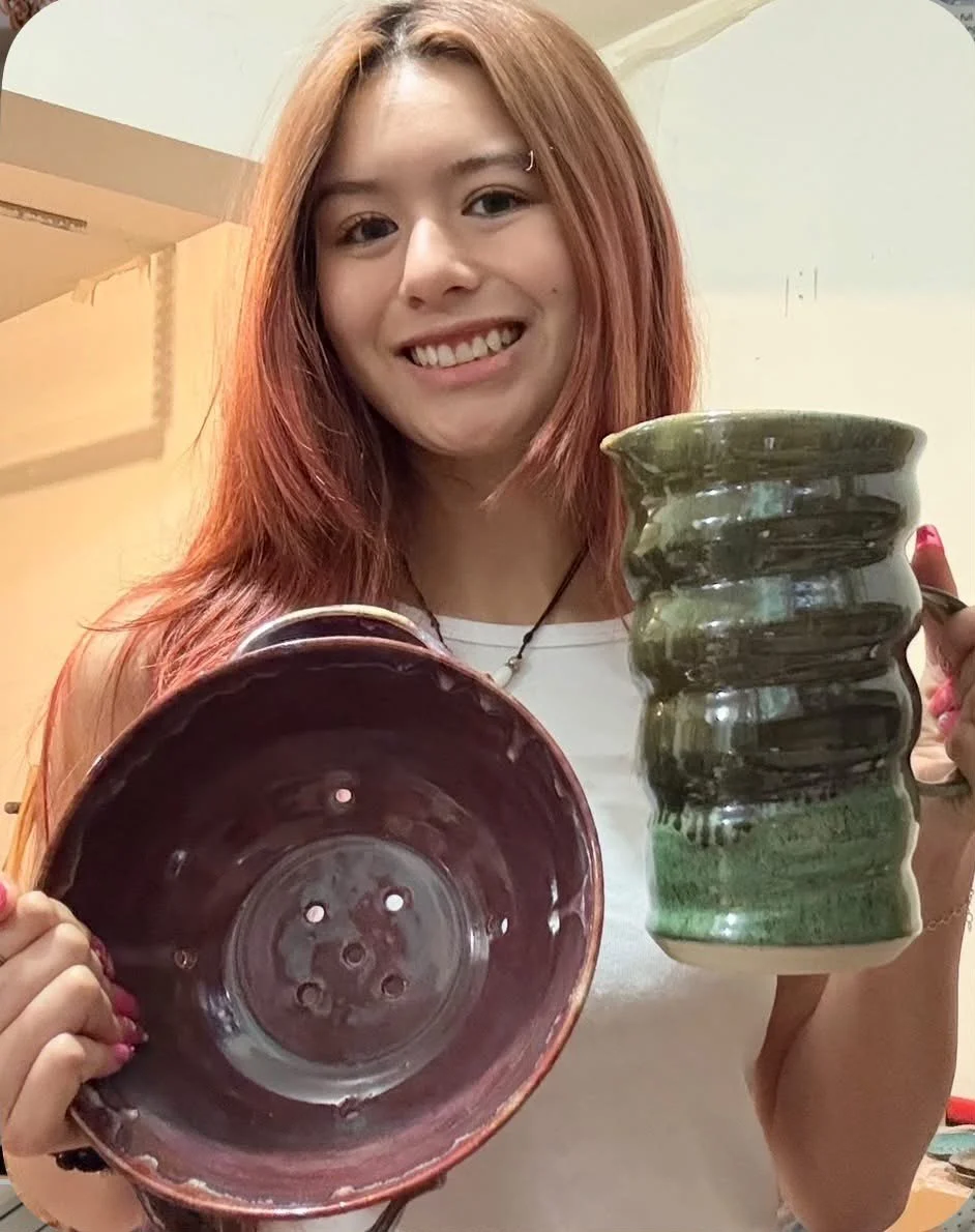 Young woman with red hair holding a green glazed pottery mug and an empty pottery bowl, smiling at the camera.