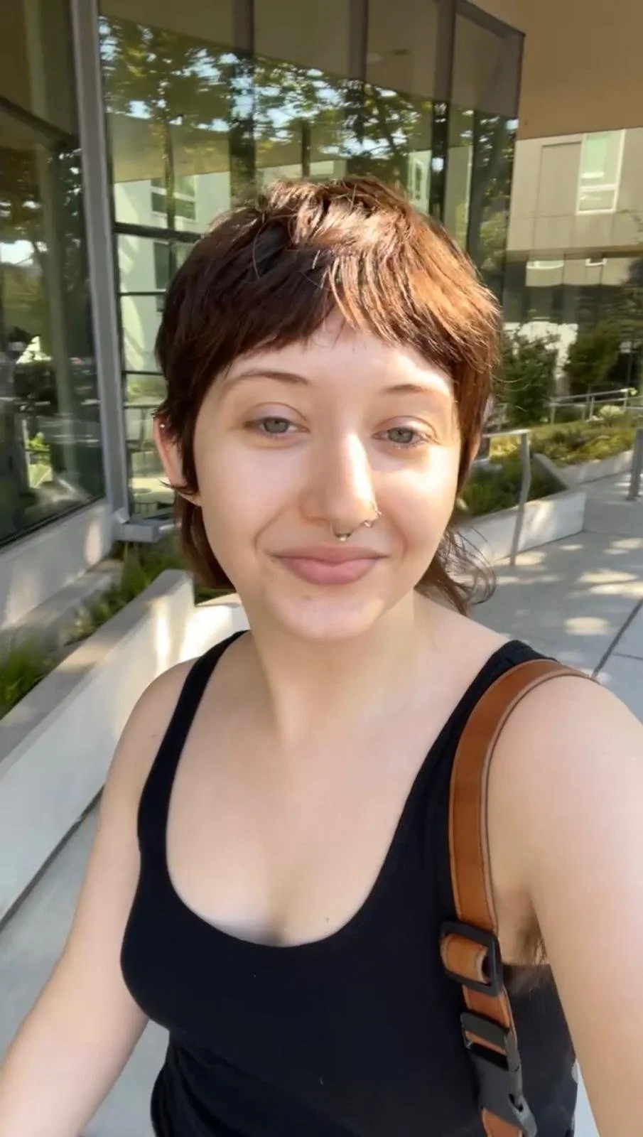 A person with short brown hair, wearing a black tank top and a brown shoulder bag, smiling outdoors with a modern building and glass windows in the background.