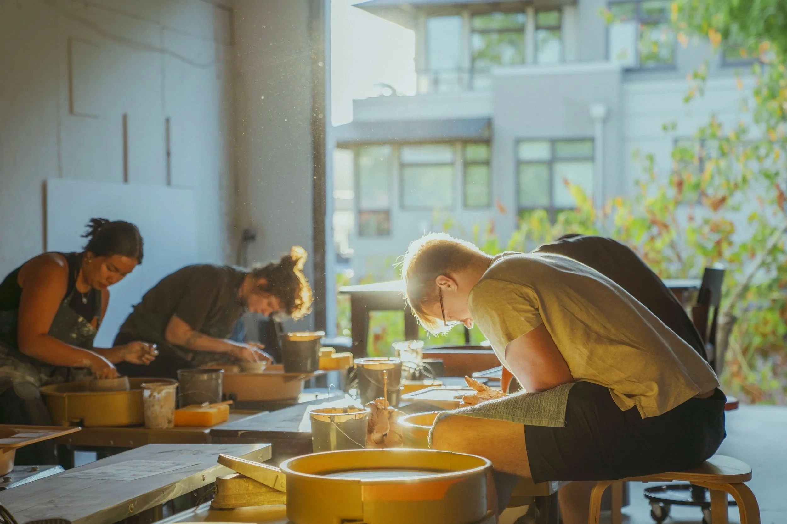 People working on pottery wheels in a sunlit workshop with large windows and greenery outside.