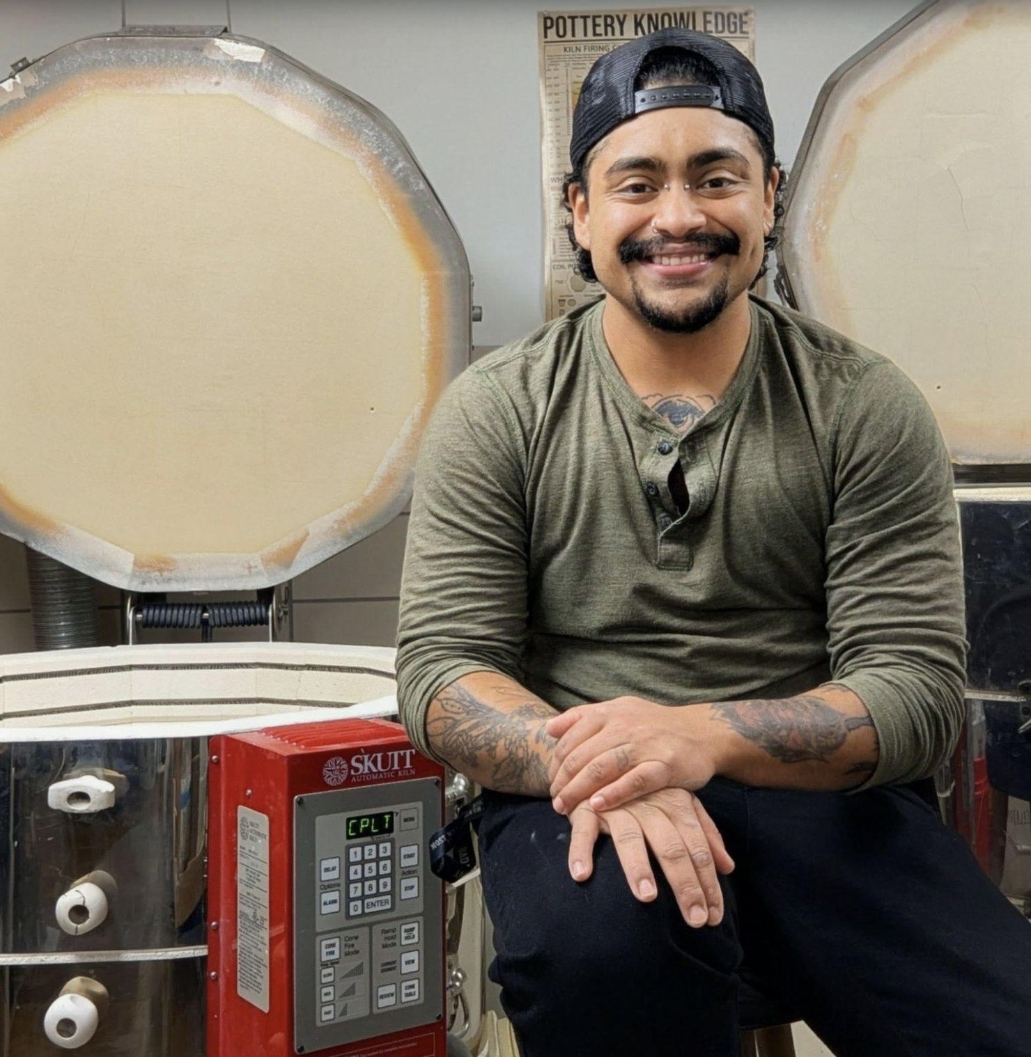 A smiling man with a mustache and tattoos on his arms, wearing a backwards black cap and a green long-sleeve shirt with a button neck, sitting in front of a kiln in a pottery studio.