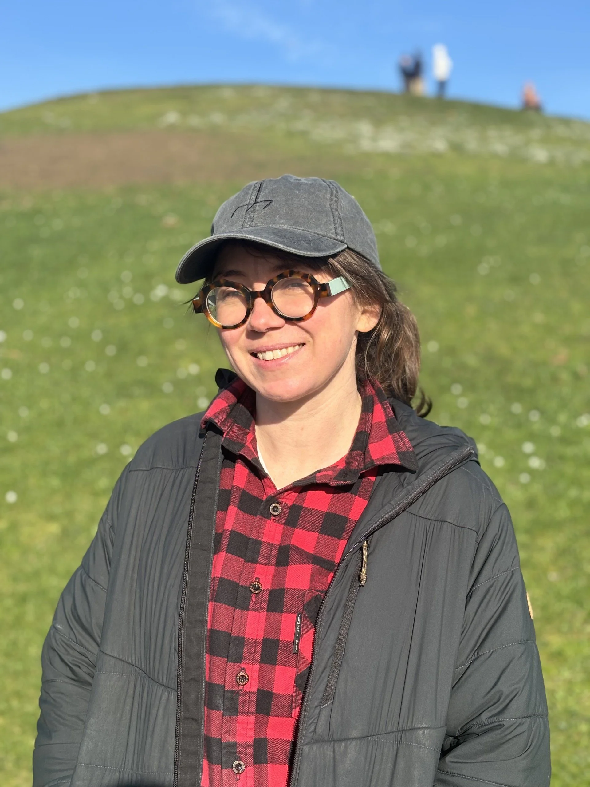 A smiling woman wearing glasses, a gray baseball cap, a red and black plaid shirt, and a gray jacket standing outdoors on a sunny day with a grassy hill and three people in the background.