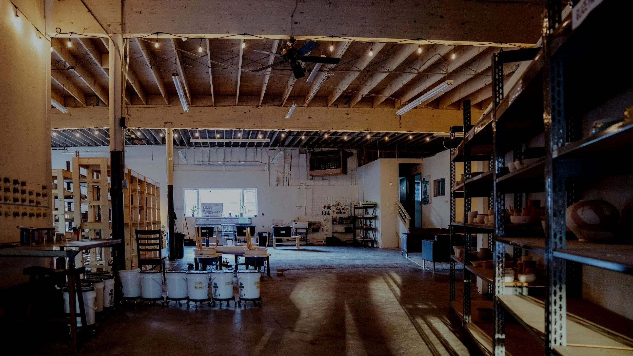 Interior of the studio with high wooden ceiling, hanging light bulbs, metal shelves on the right holding various objects, tables, chairs, and natural light coming from a large window.