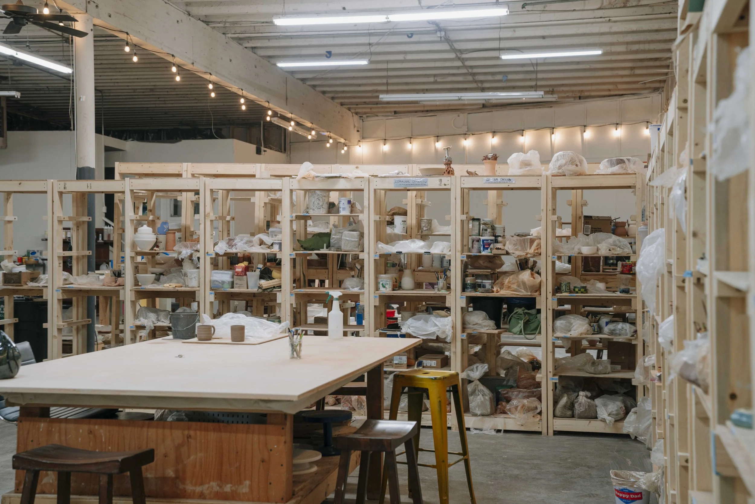 A pottery studio with wooden shelves filled with clay and ceramic art pieces, tools, and supplies; a large table with pottery in progress and stools, with a ceiling adorned with string lights and fluorescent lights.