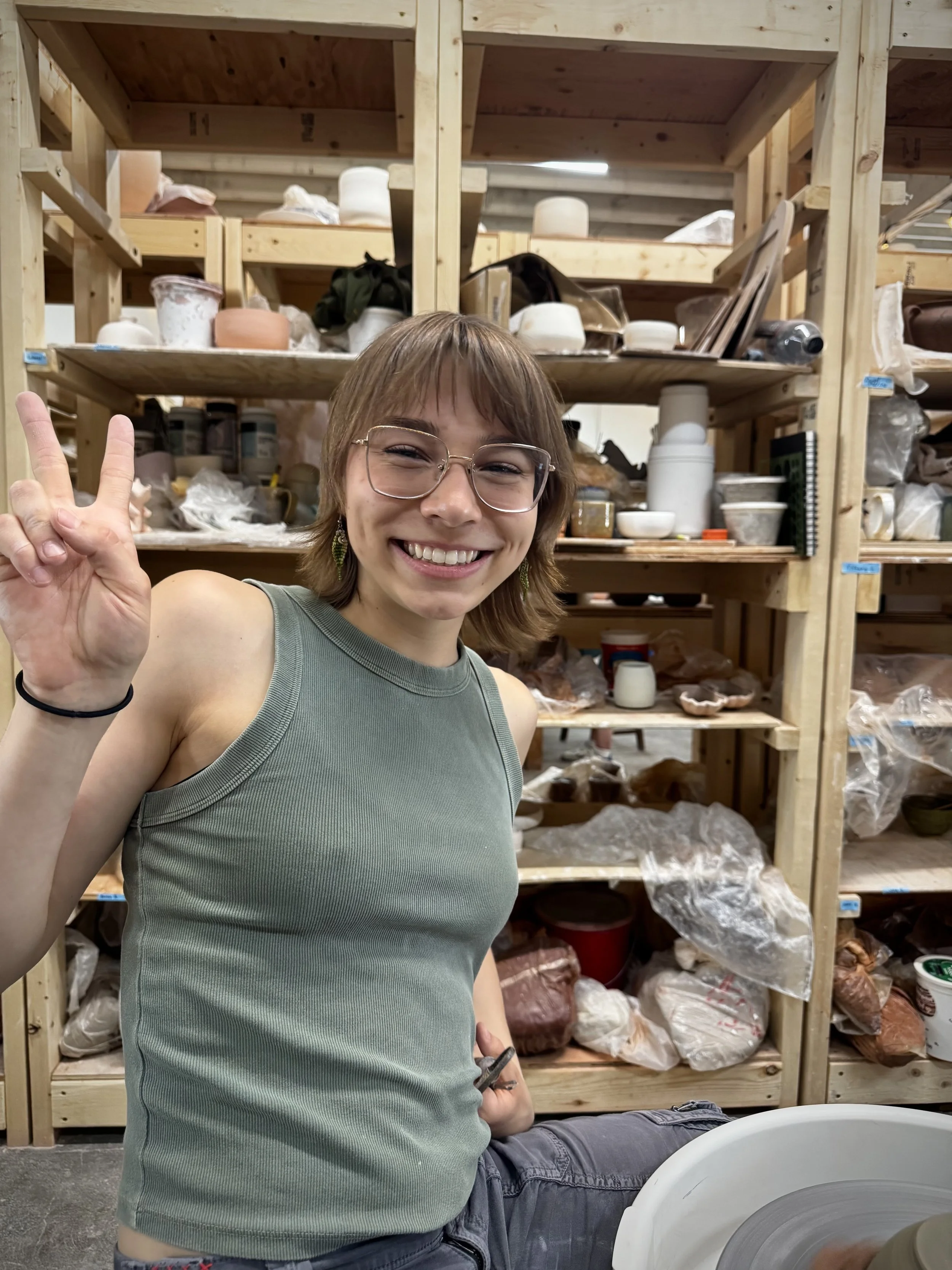 A smiling person with glasses making a peace sign in front of shelves filled with ceramic supplies in a pottery studio.