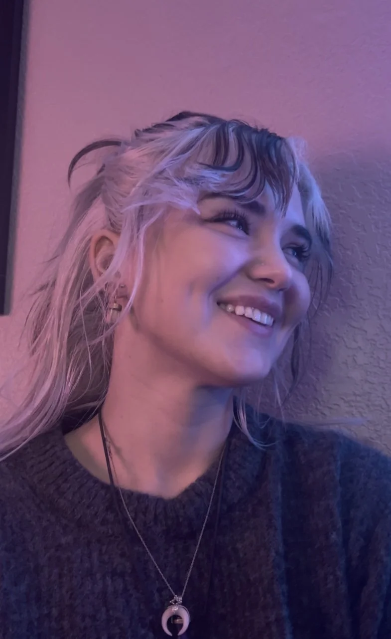 A young woman with long, wavy blonde hair, styled with dark streaks, smiling and looking to her right. She is wearing a dark sweater and a necklace with a crescent moon pendant, standing against a textured wall with purple lighting.