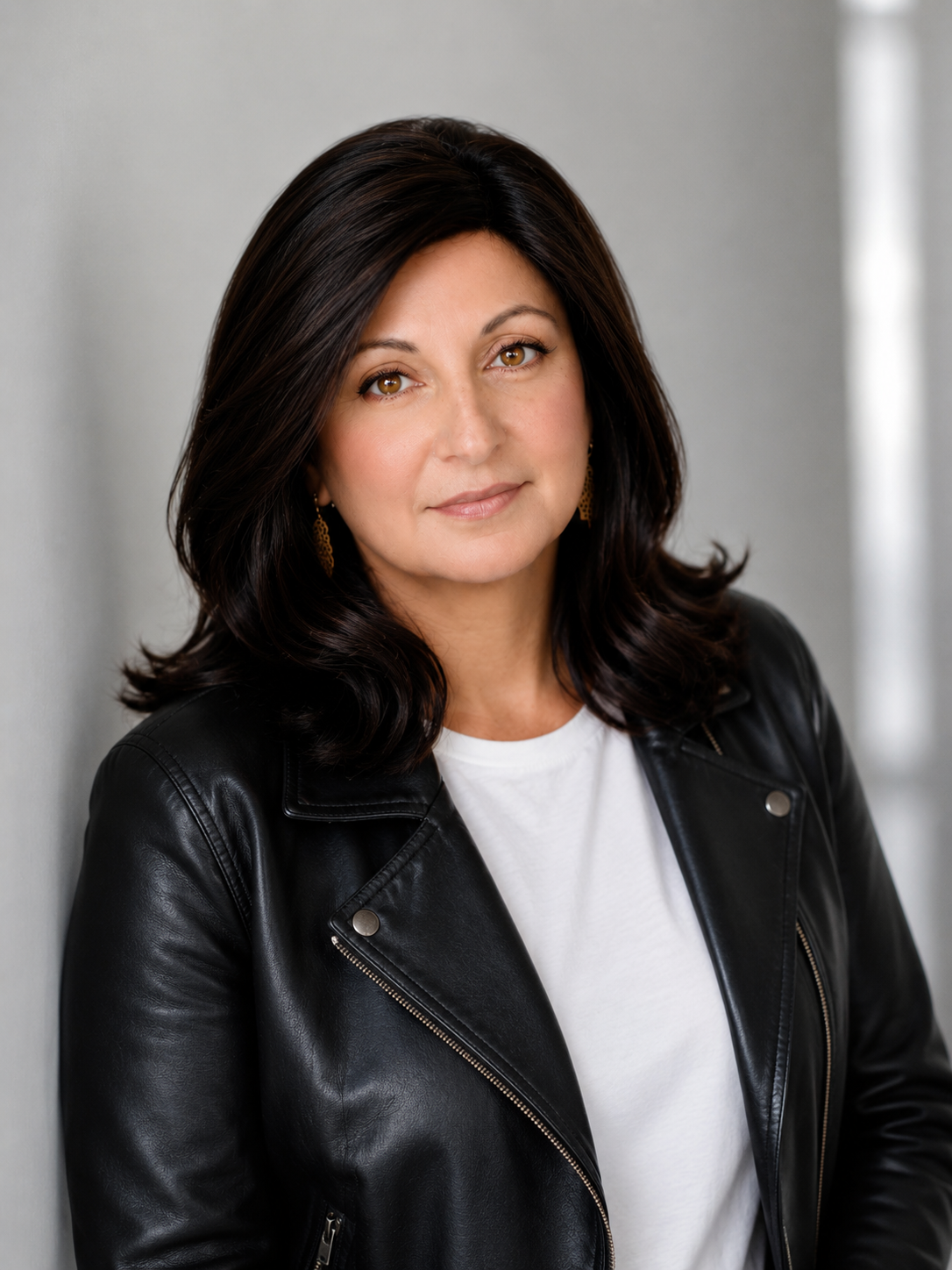 A woman with brown hair and brown eyes wearing a black leather jacket and white shirt, standing against a light gray wall.