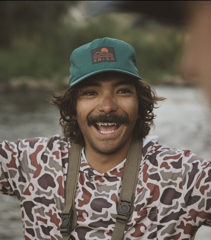 A man with curly hair, a mustache, and a beard smiling widely, wearing a teal cap with a sunset logo and the word "AMIGO," a camouflage-patterned shirt, and a camera strap around his neck, outdoors near a body of water.