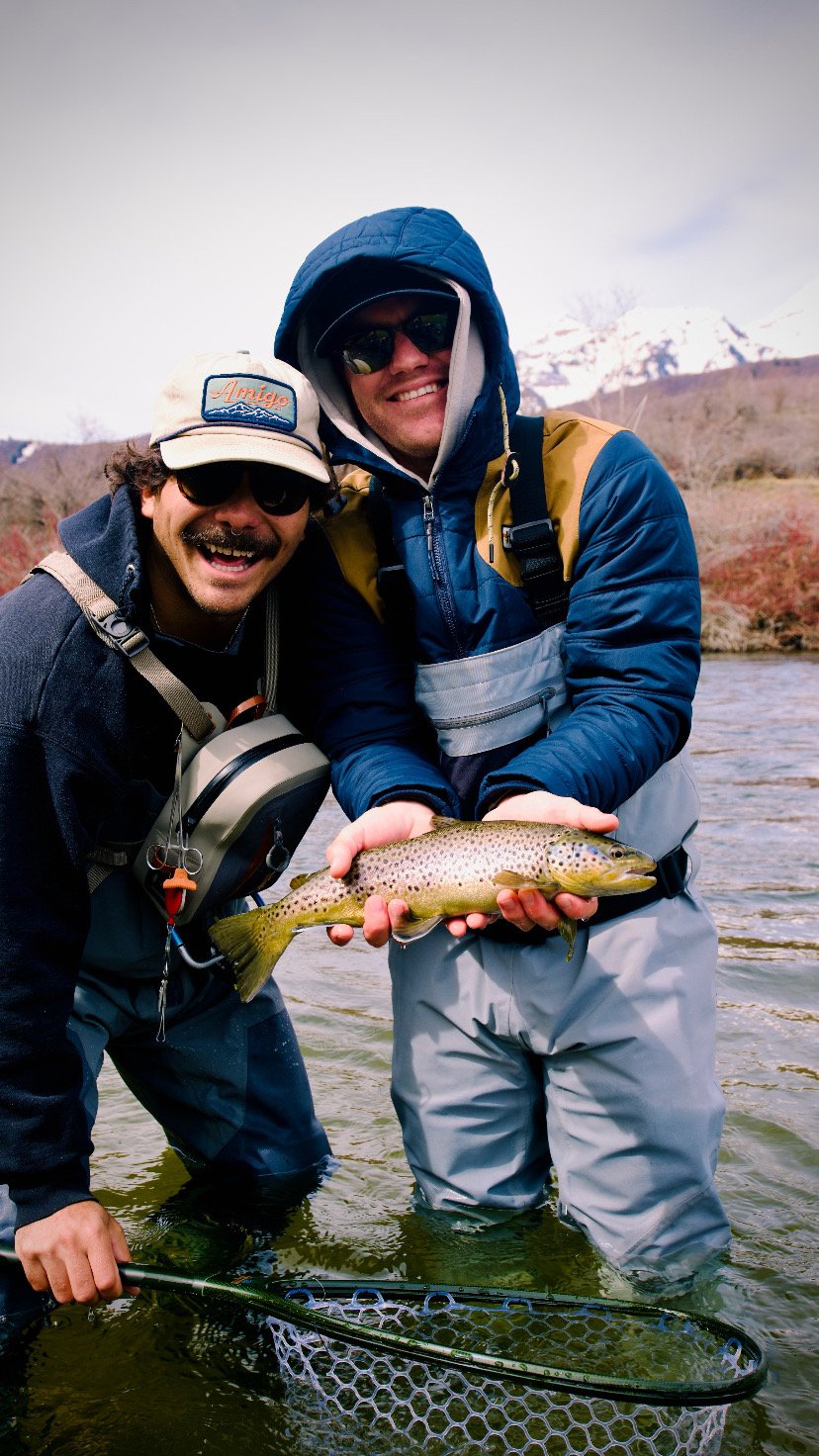 Two men in outdoor fishing gear standing in a river, holding a large fish they caught together, with a mountain landscape in the background.