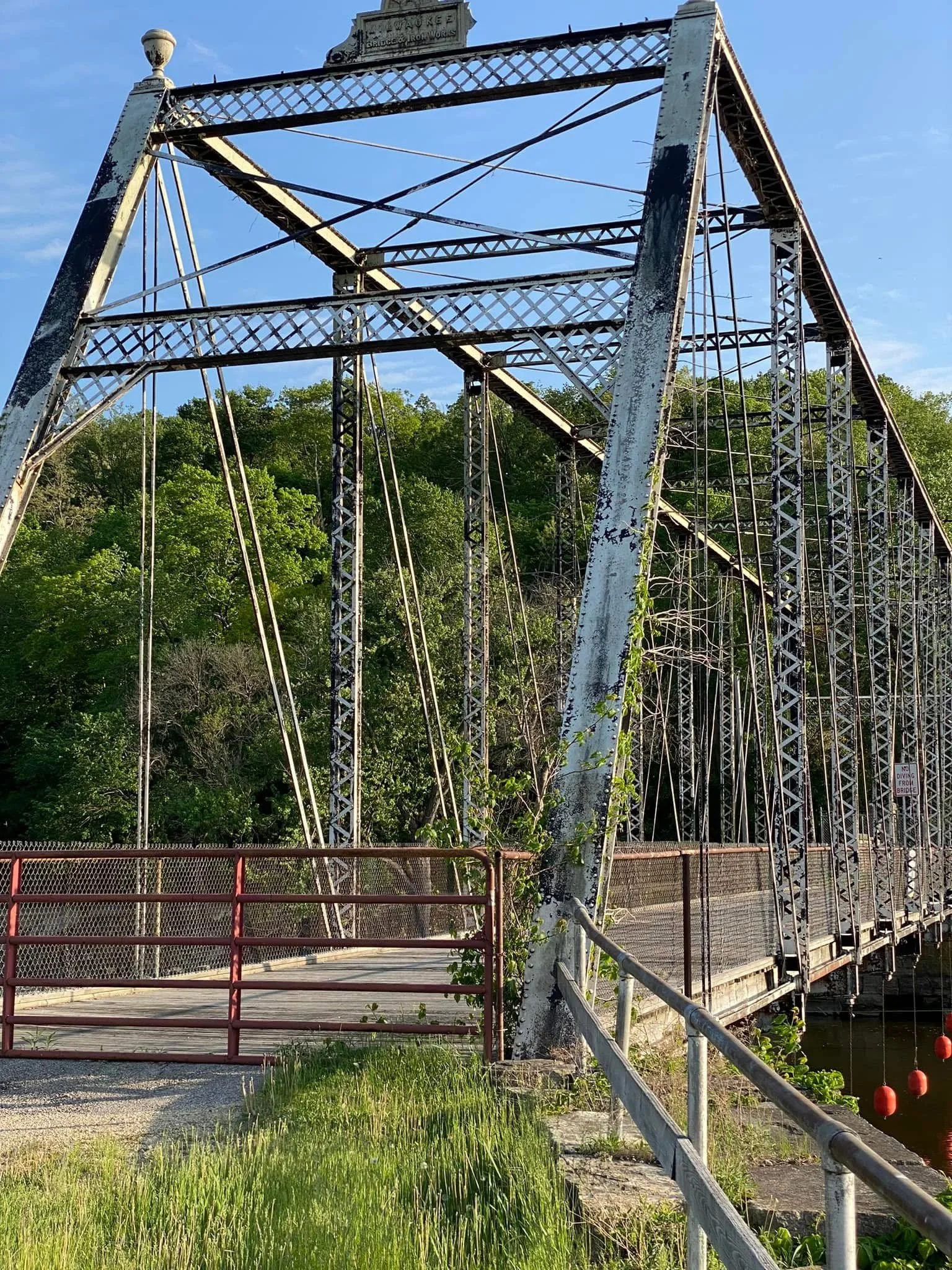 Photograph by Ashley Whitt of a historic iron truss bridge in Iowa, originally built by Milwaukee Bridge & Iron Works in 1887, featuring latticework, steel trusses, and a weathered wooden deck beneath a bright blue sky.