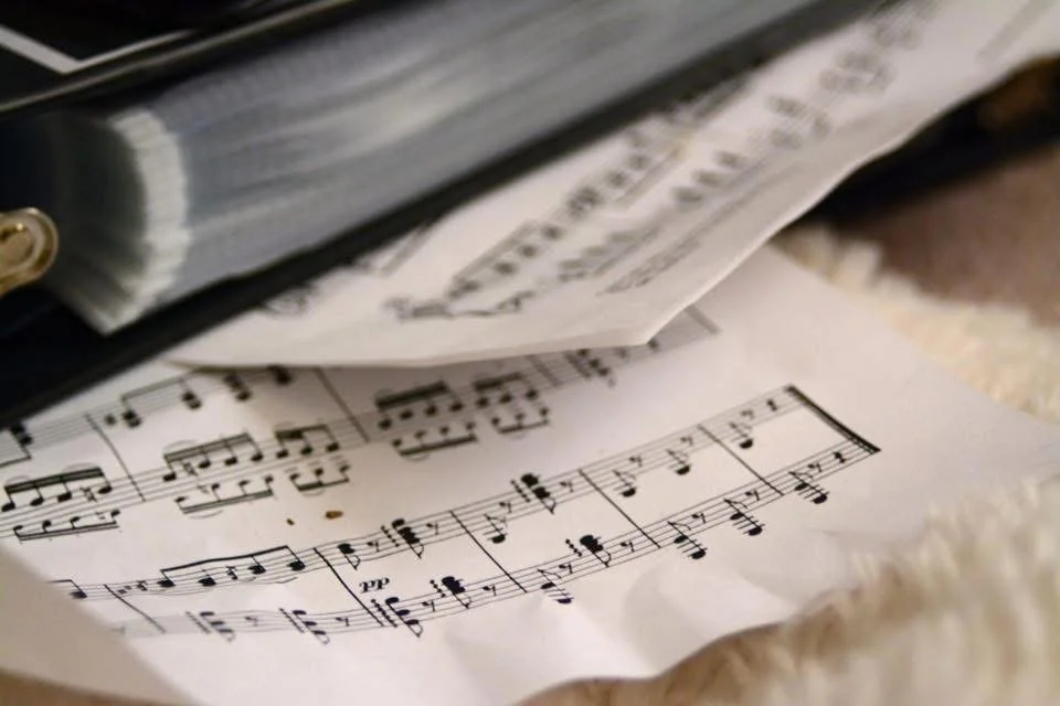 Closed binder of sheet music resting on additional music, representing Ashley Whitt's lifelong love of music and creative expression.