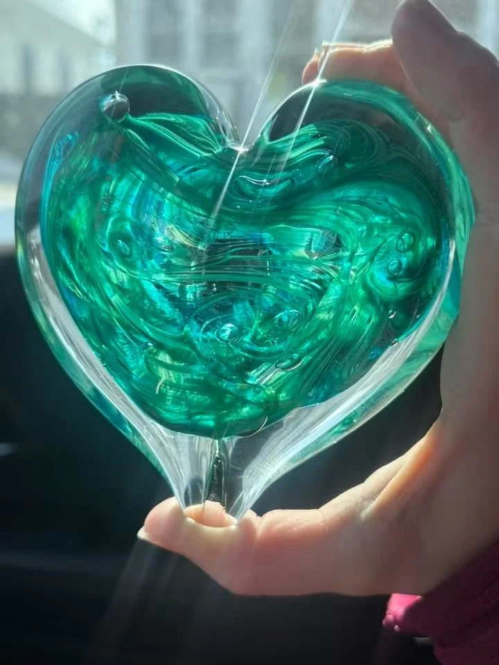 Hand holding a heart-shaped glass paperweight created by Ashley Whitt during a glassblowing class, featuring swirling shades of blue and green illuminated by natural light.