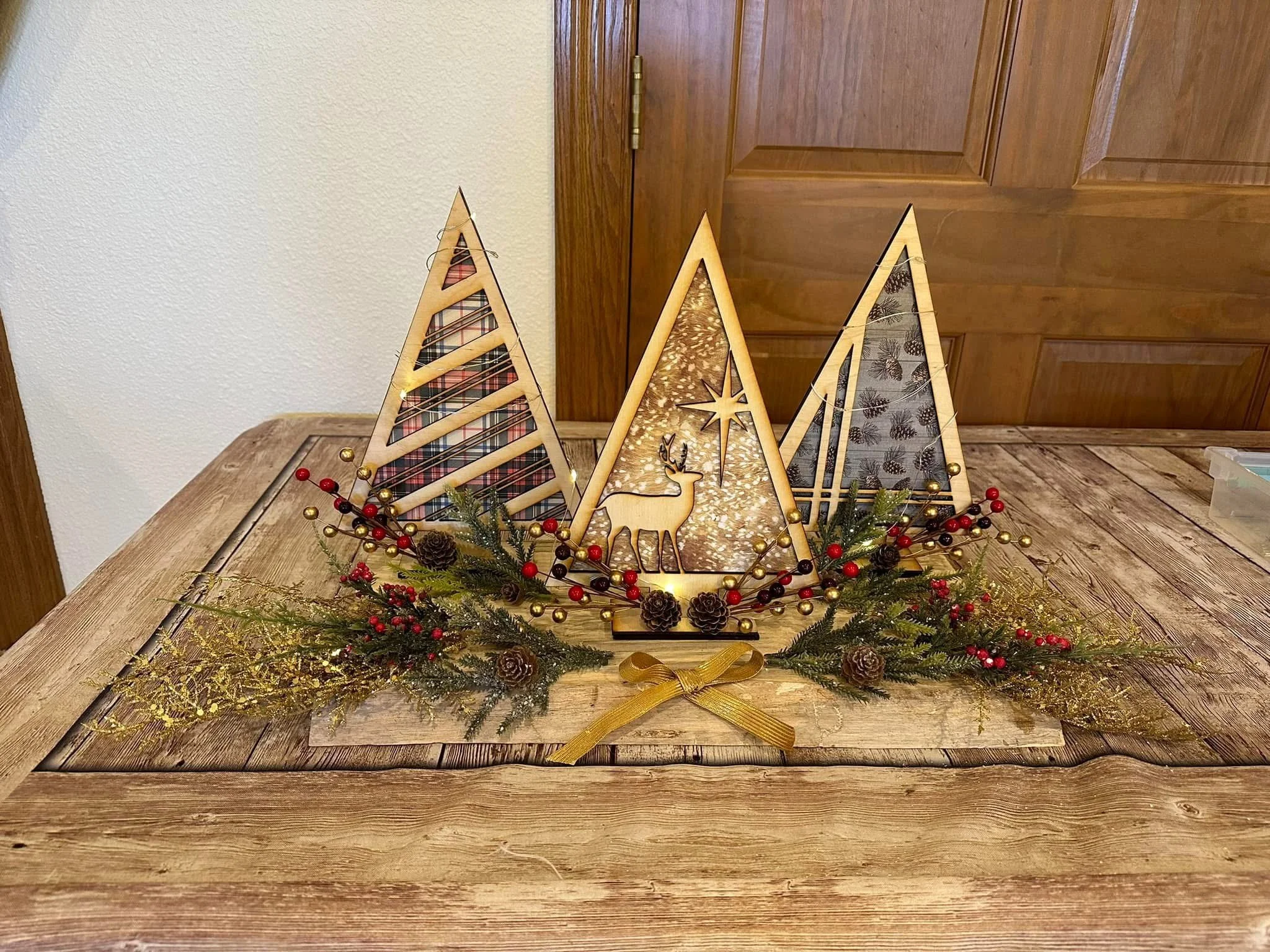 Decorative wooden Christmas trees crafted by Ashley Whitt, featuring plaid and woodland patterns, displayed with greenery, pinecones, red berries, gold ornaments, and a gold ribbon on a wooden table against a wood-paneled door.