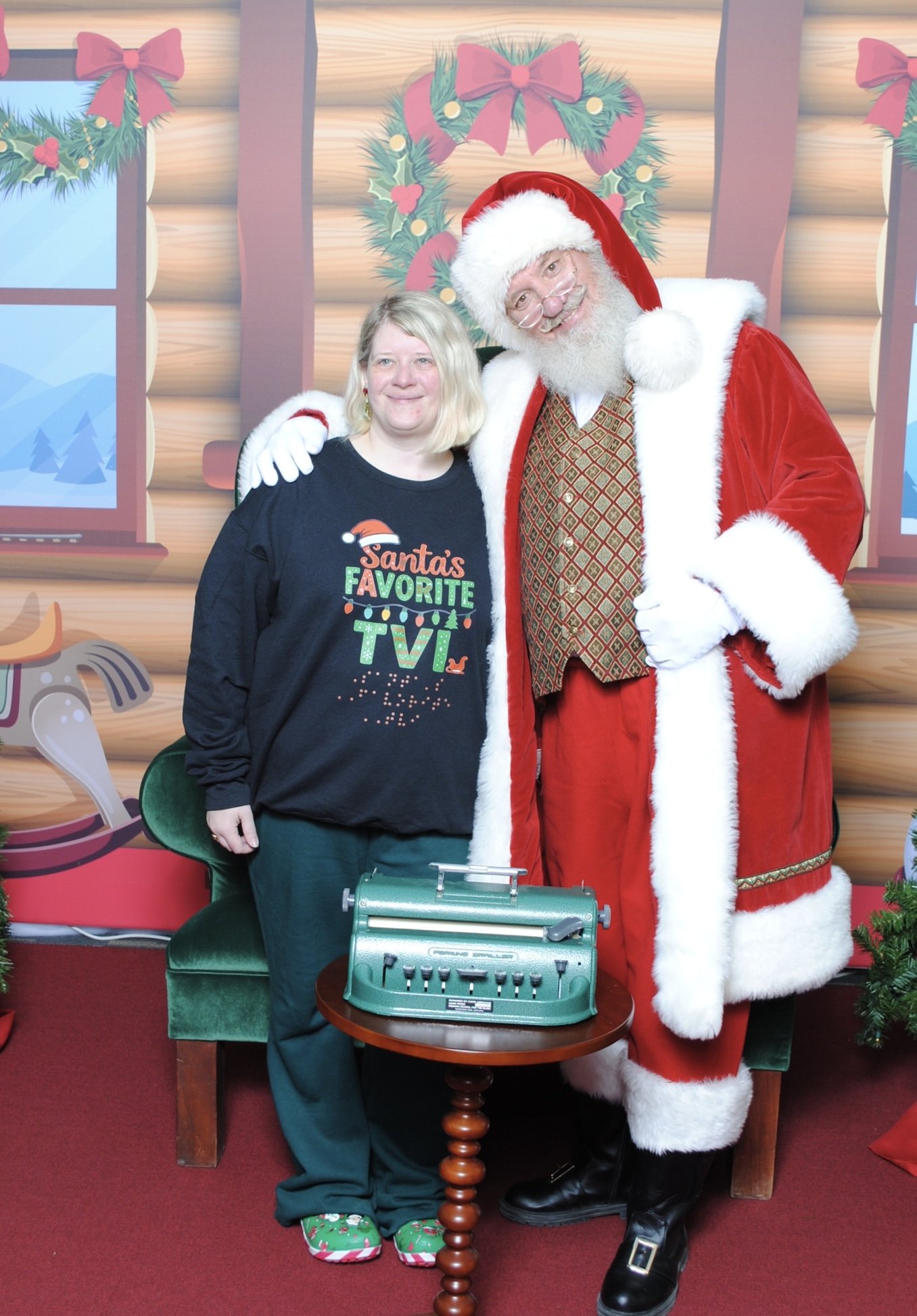 Ashley Whitt posing with Santa Claus beside a Perkins Brailler while wearing a "Santa's Favorite TVI" shirt printed in both print and braille, illustrating creative and accessible holiday learning.