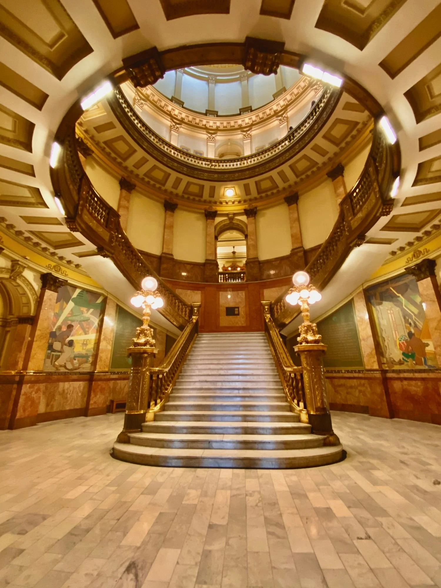 Photograph by Ashley Whitt of the grand marble staircase inside the Colorado State Capitol in Denver, Colorado, featuring ornate gold railings, chandeliers, and a domed ceiling.